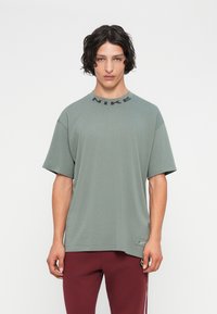 Mlad moški z valovitimi temnimi lasmi nosi preveliko majico Nike v umirjeni zeleni barvi in burgundske hlače, stoji ob svetli enostavni ozadju.