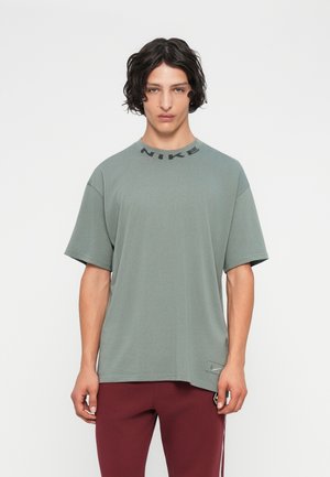 Giovane con capelli ondulati scuri indossa una maglietta Nike oversize verde pallido e pantaloni bordeaux, in piedi contro uno sfondo chiaro e neutro.