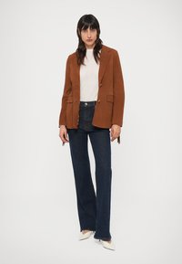 Blazer en daim marron à deux boutons, associé à une chemise gris clair et un jean évasé bleu foncé. Des chaussures blanches à bouts pointus complètent la tenue.