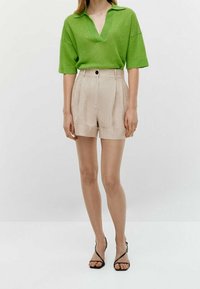 Grøn strikket polo shirt med korte ærmer, parret med beige skræddersyede shorts med høj talje, der har en enkelt knap og oprullede manchetter. Tynde sorte sandaler.