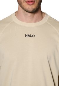 Beżowa bluza z okrągłym dekoltem, rękawami raglanowymi i wyszytym na czarno napisem "HALO" na klatce piersiowej. Gładka, miękka tkanina.
