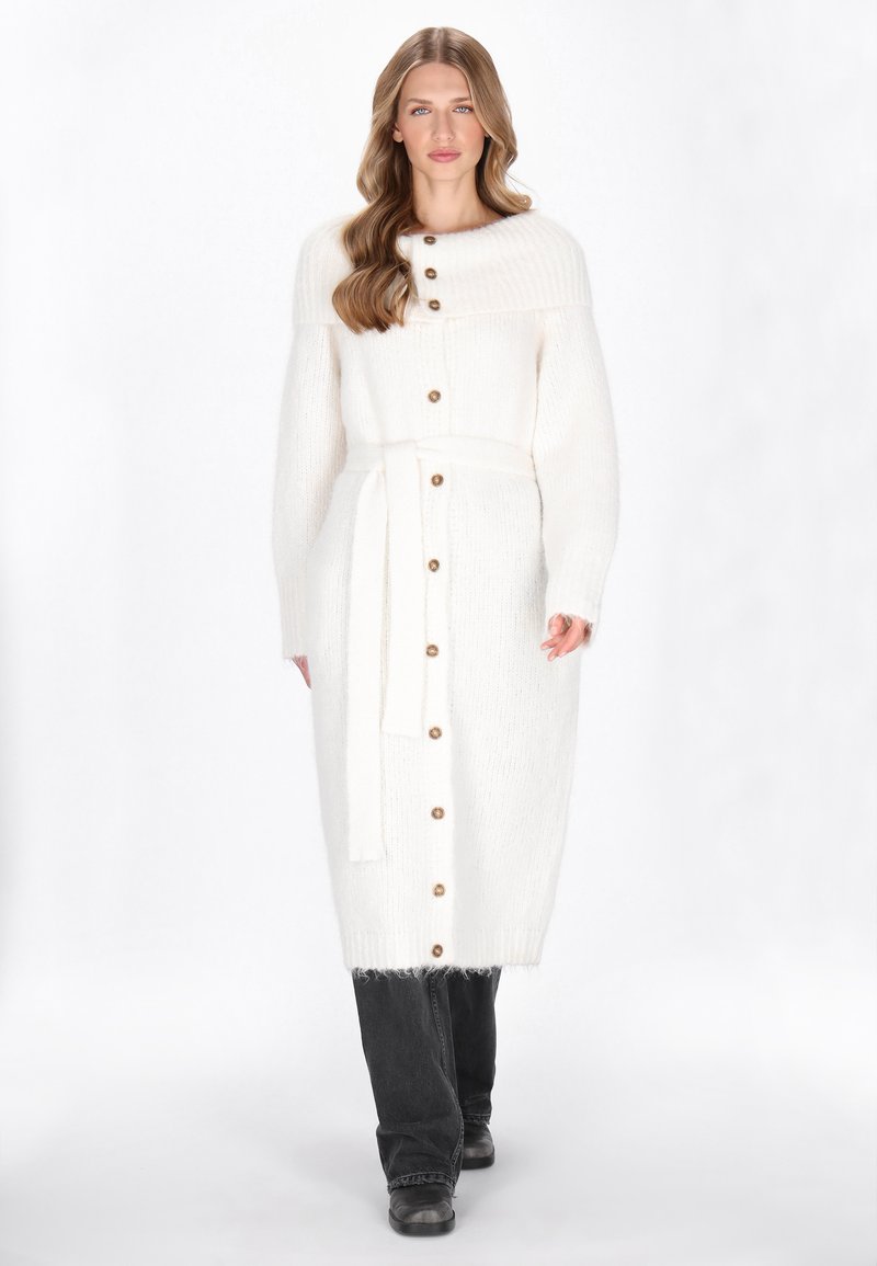 Lange witte gebreide cardigan met een geribde kraag, knoopsluitingen, een tailleband en zijzakken, gecombineerd met zwarte jeans en laarzen.