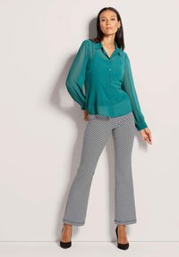 Teal-blaues Blusenhemd mit transparenten Ärmeln, kombiniert mit schwarzen und weißen, gezackten, ausgestellten Hosen. Schwarze High Heels runden das Outfit ab.