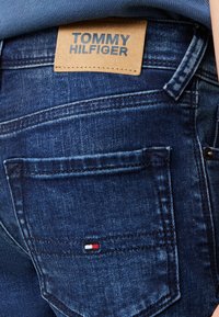 Jeans in denim scuro con un'etichetta in pelle visibile con il testo "TOMMY HILFIGER". La tasca posteriore presenta un piccolo accento del logo rosso, bianco e blu.