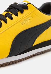 Žlutočerné tenisky Puma s texturovaným syntetickým svrškem, zavazováním na tkaničky a gumovou podrážkou. Obsahují značky "PUMA" a "ROMA".