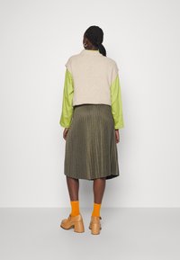 Gilet beige a maglia sopra una camicia verde brillante, abbinato a una gonna plissettata olivastro e scarpe con tacco chunky color sabbia, arricchito da calze arancioni.