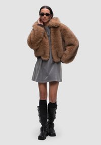 Giacca corta in pelliccia marrone con una texture morbida, vestito grigio sotto e stivali neri al ginocchio con cinturini e fibbie.