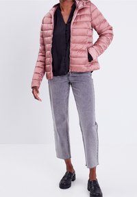 Chaqueta acolchada con capucha en rosa claro con un acabado brillante, cierre de cremallera y bolsillos laterales, combinada con jeans grises y zapatos de plataforma negros.