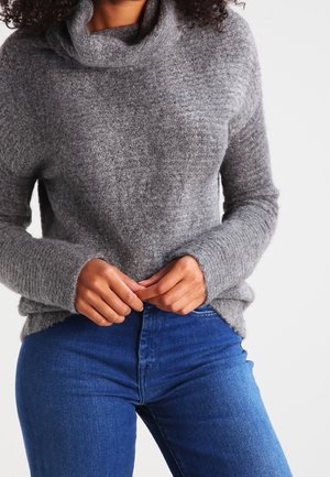 Personne portant un pull gris texturé à col boule et un jean bleu, les mains jointes près de la taille sur un fond uni.