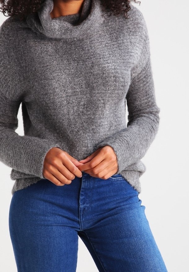 Personne portant un pull gris texturé à col boule et un jean bleu, les mains jointes près de la taille sur un fond uni.