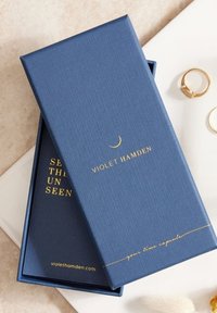 Navy blue textured box with gold text. Inner box features "SEE THE UNSEEN" in gold. Includes a golden crescent moon accent.