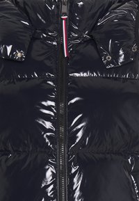 Glansig svart pufferjacka med central dragkedja och knappknäppning vid ärmsluten, med en dragkedjetab som är röd, vit och blå.