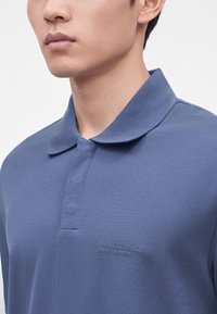 Νεαρός άνδρας που φοράει μπλε polo Armani Exchange με μαλακό γιακά, εμφανιζόμενος από το στήθος και πάνω σε απλό φόντο.
