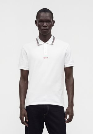 Homme portant un polo blanc Versace avec un col à motifs et un pantalon noir sur un fond blanc uni.
