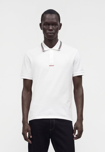 Homme portant un polo blanc Versace avec un col à motifs et un pantalon noir sur un fond blanc uni.
