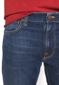 Lähekuva henkilöstä, joka käyttää tummansiniset farkut oranssilla ompelulla ja punaiseksi paidan, joka on taltioitu vyötärölle.