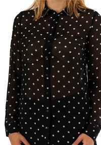Woman wearing a black sheer blouse with white polka dots and a studded collar, buttons down front, and long sleeves.