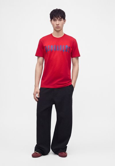 Jeune homme portant un T-shirt rouge Dsquared2, un pantalon noir ample et des chaussures rouges, debout devant un fond blanc uni.