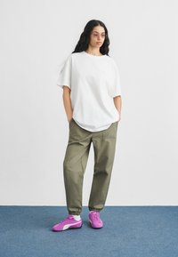 Γυναίκα στέκεται με τα χέρια στις τσέπες, φοράει λευκό φαρδύ t-shirt, χακί παντελόνι jogging, μωβ αθλητικά παπούτσια και γυαλιά με ροζ απόχρωση, σε μπλε πάτωμα.