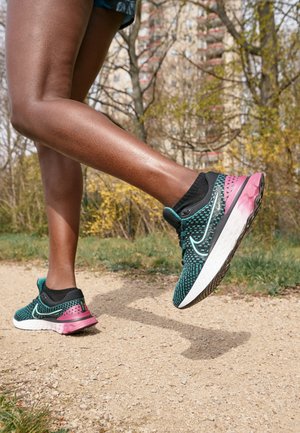 Jambes en chaussures de course bleu sarcelle et rose marchant sur un chemin en terre avec des arbres et un bâtiment en arrière-plan.