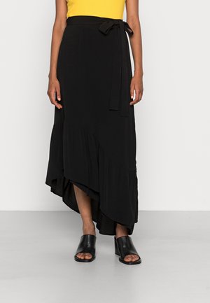 Femme portant une jupe portefeuille asymétrique noire avec une ceinture nouée, associée à des mules à talons ouvertes noires et un top jaune.
