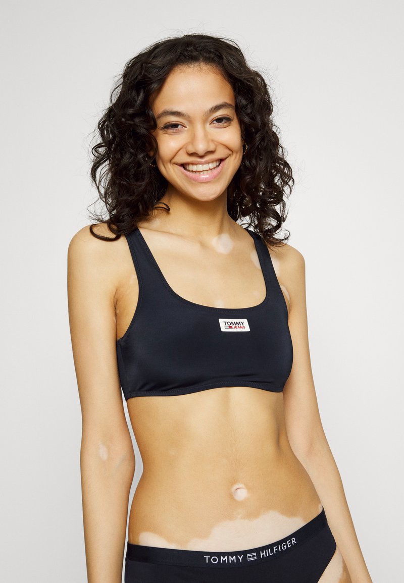 Sujetador deportivo negro con tirantes anchos, que presenta un pequeño logo de Tommy Jeans en rojo y blanco. Textura suave, diseño minimalista. Cinturilla negra por debajo.