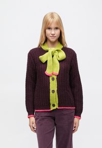 Megztinis tamsiai vyno raudonos spalvos, turintis ryškiai žalių sagų juostelę, rožinius akcentus, tekstūruotą audinį ir didelę lanką prie kaklo.