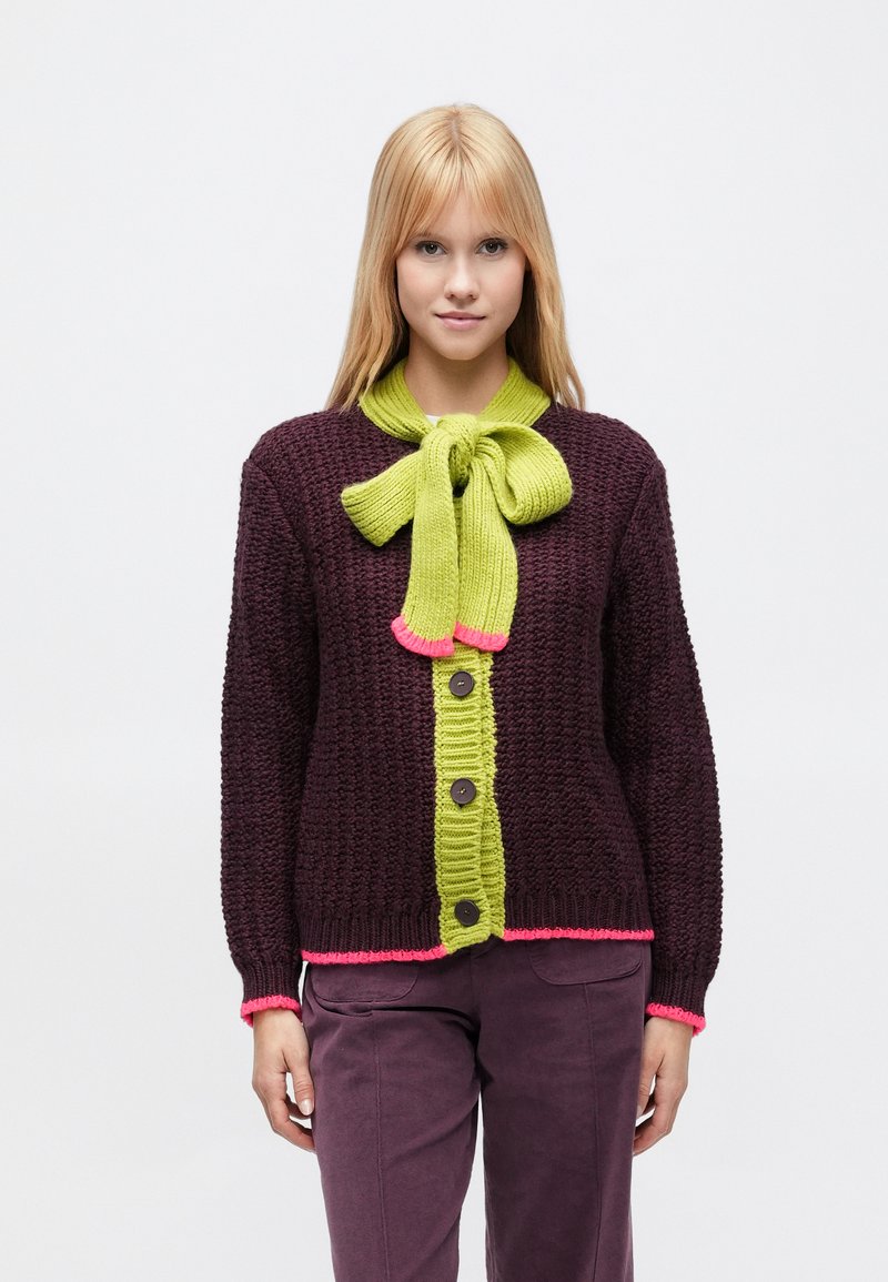 Megztinis tamsiai vyno raudonos spalvos, turintis ryškiai žalių sagų juostelę, rožinius akcentus, tekstūruotą audinį ir didelę lanką prie kaklo.