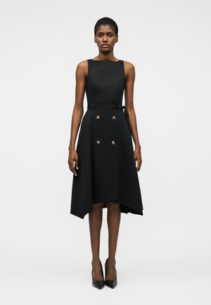 Femeie purtând o rochie midi neagră fără mâneci, cu tiv asimetric, nasturi cu două rânduri și curea, combinată cu pantofi negri cu toc ascuțit, stând drept.