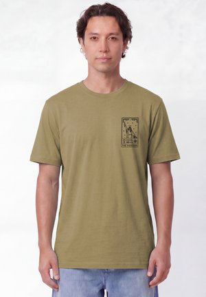 Homme portant un T-shirt vert olive avec un petit motif de carte de tarot noire étiqueté "The Magician" sur la poitrine gauche, associé à un jean bleu clair.
