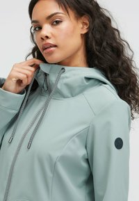 Light green waterproof jacket with a hood, featuring adjustable drawstrings and a round black logo patch on the sleeve. Smooth texture.