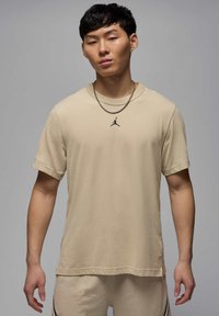 Beige kortærmet T-shirt i et blødt stof med en rund halsudskæring og et lille sort Jordan-logo på brystet. Bæres med lyse shorts.