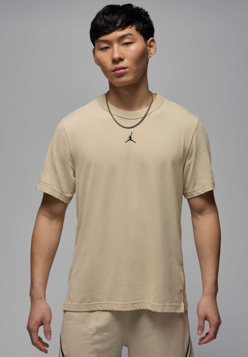 Beige kortærmet T-shirt i et blødt stof med en rund halsudskæring og et lille sort Jordan-logo på brystet. Bæres med lyse shorts.