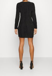 Černé šaty s dlouhým rukávem, vyrobené z texturované látky, s přiléhavým střihem a jemným nařasením, doplněné černými lodičkami na vysokém podpatku.