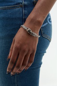 Zilveren schakelarmband met een platte, rechthoekige sluiting en gladde schakels, gedragen op een hand tegen blauwe denim. Donkere nagellak accentueert.