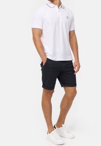 Polo blanc avec un bord de col bordeaux et un logo, associé à un short noir et des baskets blanches, avec un design à l'ourlet roulé.