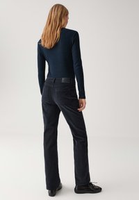 Vrouw met schouderlang blond haar, gekleed in een zwart geribbeld shirt met lange mouwen, zwarte rechte broek en zwarte schoenen, staande met de rug naar de kijker gekeerd.