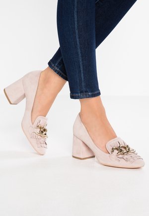 Mujer con zapatos de tacón cuadrado de ante rosa claro, con cadena dorada y detalle de flecos, combinados con vaqueros ajustados azul oscuro.