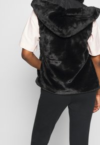 Black faux fur vest with a hood, featuring a plush texture. Worn over a light pink short-sleeve top and black pants. Simple design.