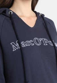 Granatowy sweter z dekoltem w serek, z kapturem na sznurku i białym haftowanym logo "Marc O'Polo" z przodu. Miękka tekstura.