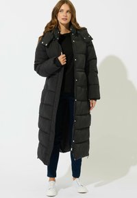 Kvinde iført en lang sort puffet frakke over en sort top og mørke bukser, der står med hvide sneakers mod en ensartet lys baggrund.