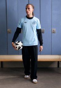 Lyseblåt træningsskjorte med sorte lange ærmer, sorte bukser og grå sportssko, som holder en sort-hvid fodbold foran skabe.