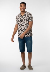 Kurzarmhemd in Beige mit schwarzen abstrakten Mustern, kombiniert mit blauen Jeansshorts und grauen Slippern. Einfaches Design und lockere Passform.