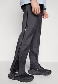 Men's black waterproof pants with side zippers, made from textured fabric, paired with black athletic shoes featuring a rugged sole.