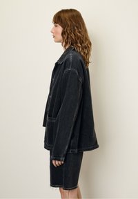Zwarte denimjack met een losse pasvorm, grote zakken en contrasterende stiksels, gecombineerd met een bijpassende denimrok. De textuur lijkt zacht en versleten.