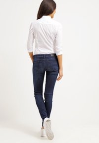 Jeans skinny azul oscuro con una textura suave, diseño de cinco bolsillos y una cintura baja, combinados con una camisa blanca con cuello y zapatillas blancas.
