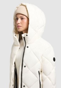 Chaqueta acolchada blanca con una gran capucha, cierre frontal negro con cremallera y detalles de botones a presión. Llevada sobre una blusa ligera, acompañada de un gorro de punto beige.