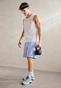Camiseta atlética sin mangas blanca, pantalones cortos púrpura claro, kettlebell negro, calcetines verde menta, zapatos grises con detalles negros; logo de Nike visible.