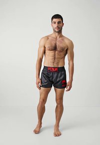 Muscularer Mann, der barfuß auf einem schlichten hellen Hintergrund steht und schwarze Venum-Shorts mit rotem Logo trägt.