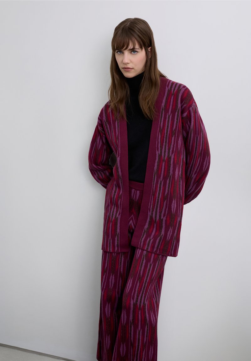 Knitted maroon cardigan and pants featuring geometric patterns in red and pink hues, with a black turtleneck underneath. Soft texture, relaxed fit.